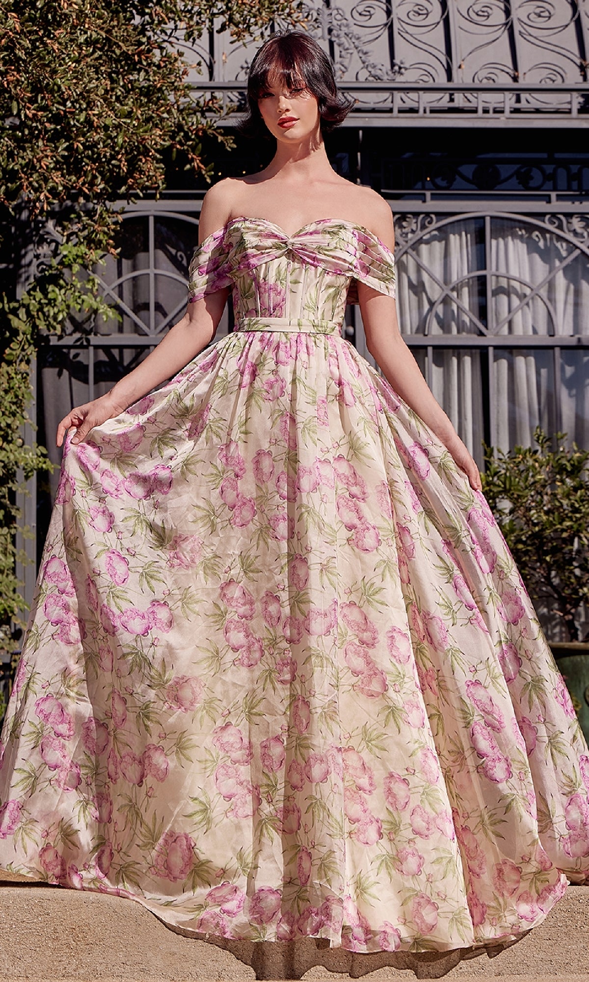 A woman stands outside by a wrought iron gate, wearing the Andrea & Leo A1466 long floral-print prom dress with an off-the-shoulder corset bodice, holding the sides of its floor-length skirt.